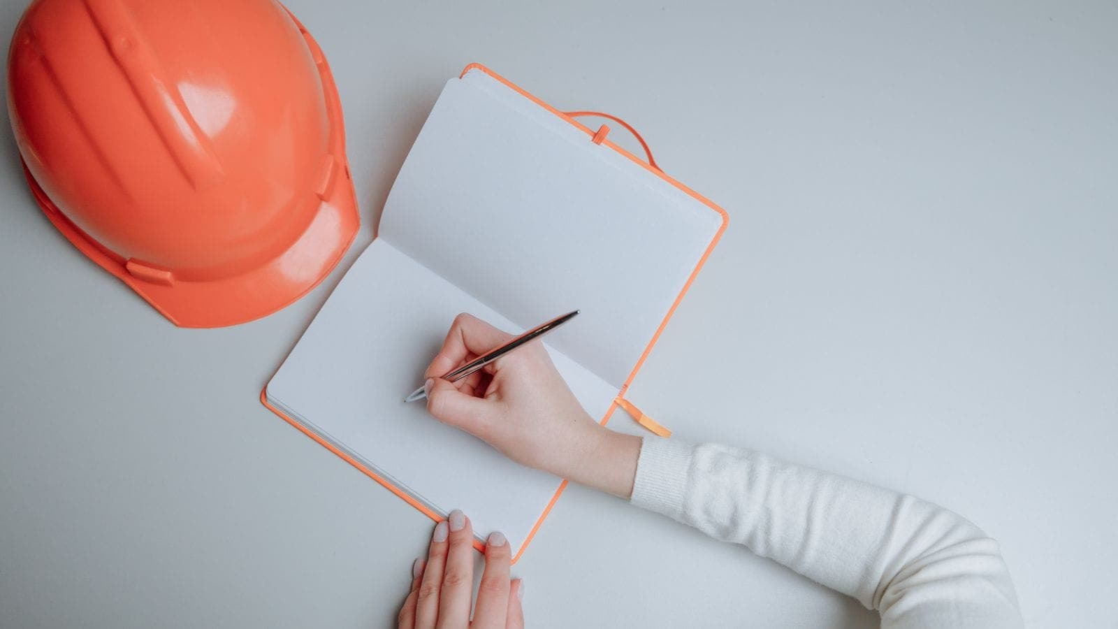 Top-down view of a person writing in a planning notebook beside an orange construction hardhat
