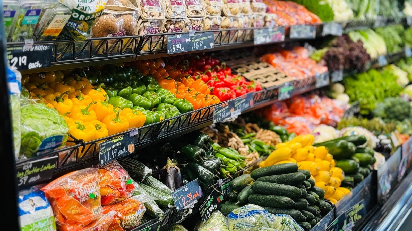 Vibrant grocery store produce section with colorful bell peppers, cucumbers, squash, and packaged greens on display shelves