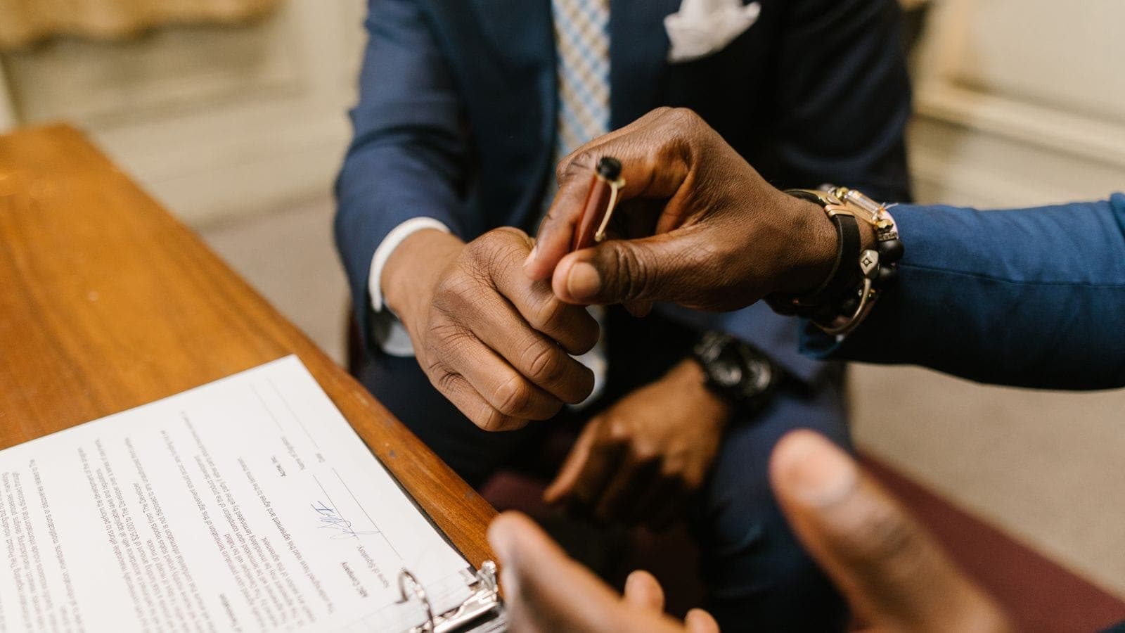 Two professionals in suits at a table, one handing a pen to the other to sign contract documents