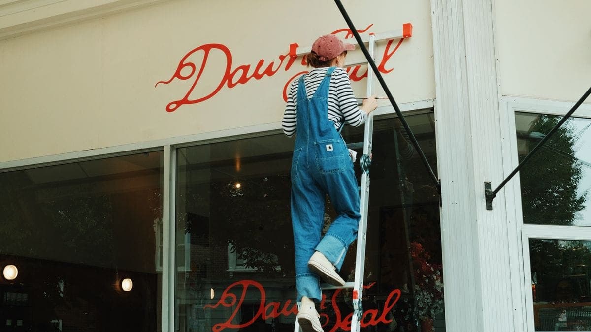 A worker in denim overalls on a ladder hand-painting a cursive sign on a cafe storefront