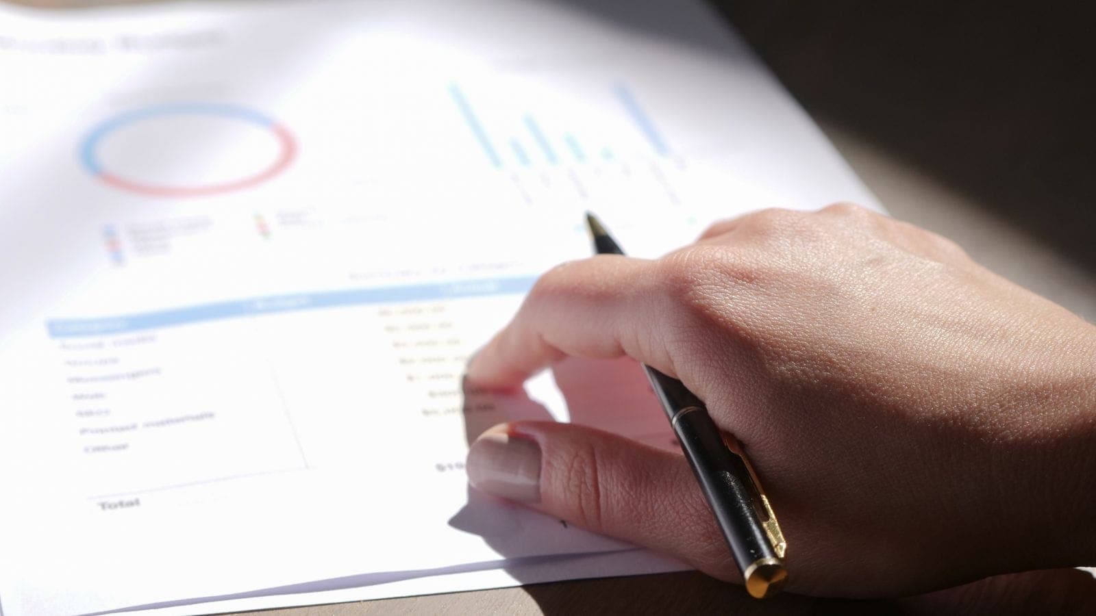 Hand holding a pen reviewing printed financial charts and budget figures on a desk