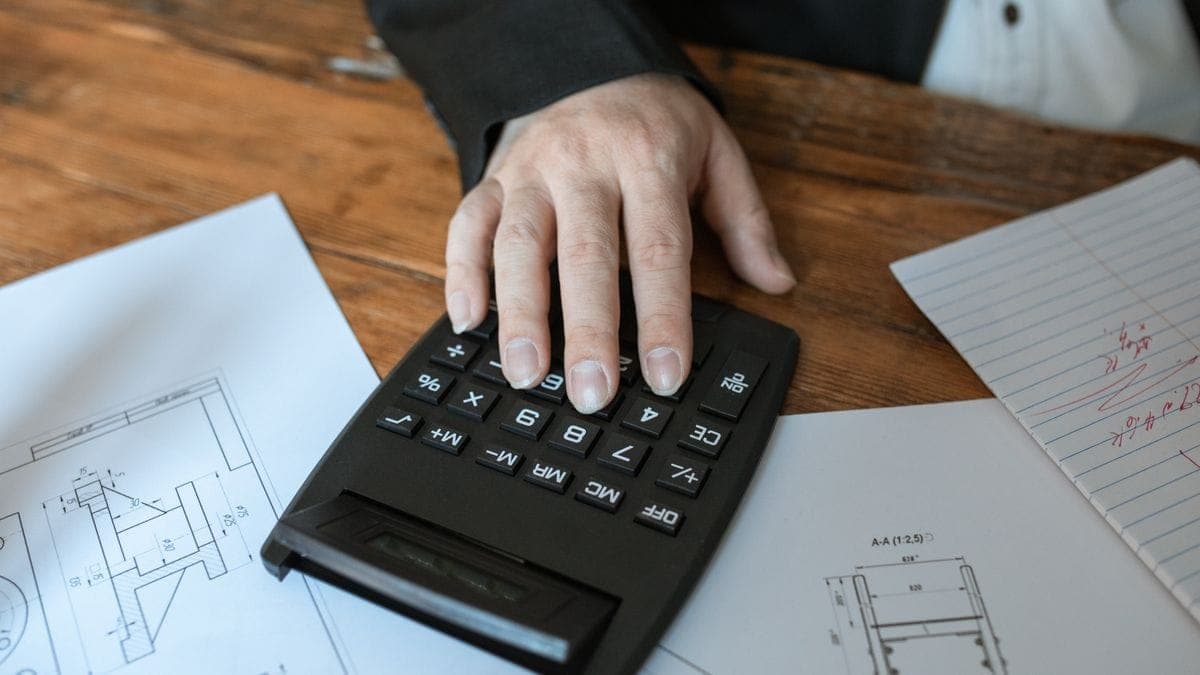 A hand using a black calculator resting on construction blueprints on a wooden desk