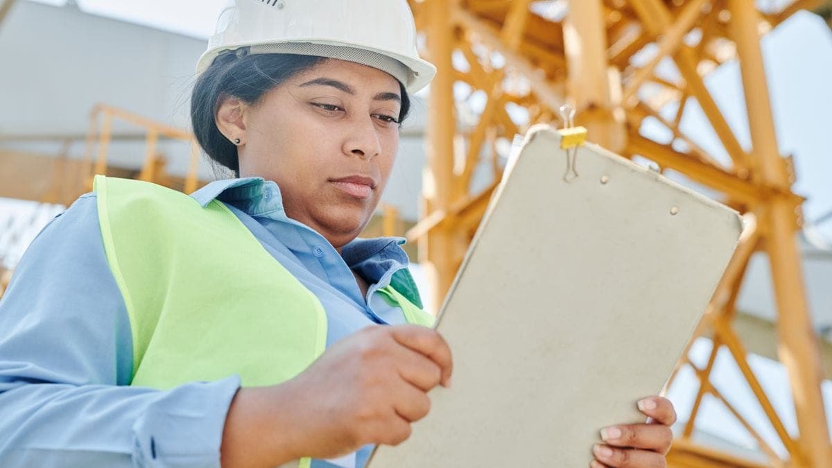 A construction worker in a hard hat and safety vest holding a clipboard while inspecting a job site